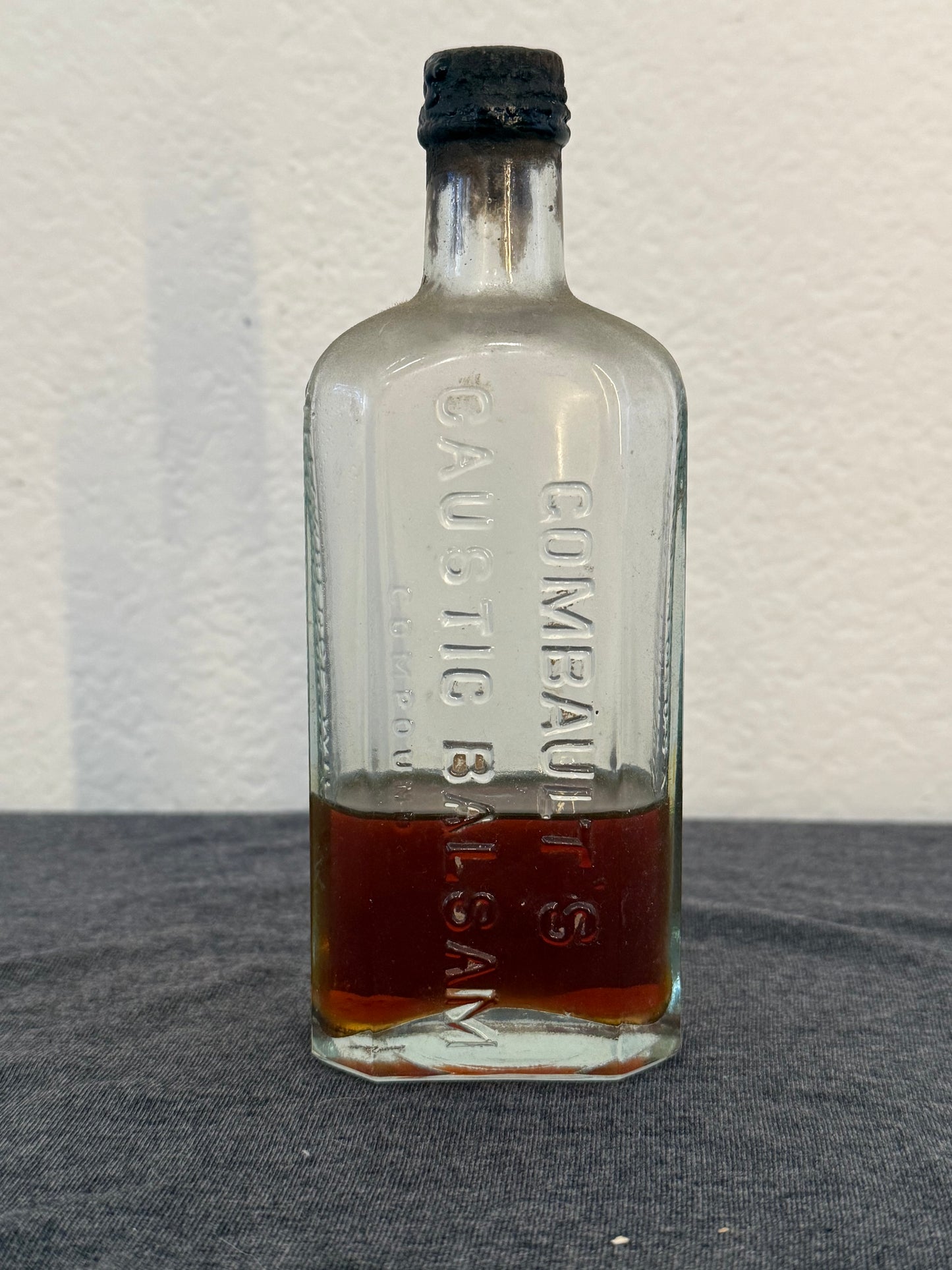 Clear glass bottle labeled 'Gumbault's Caustic Balsam' with dark liquid inside on a gray background.