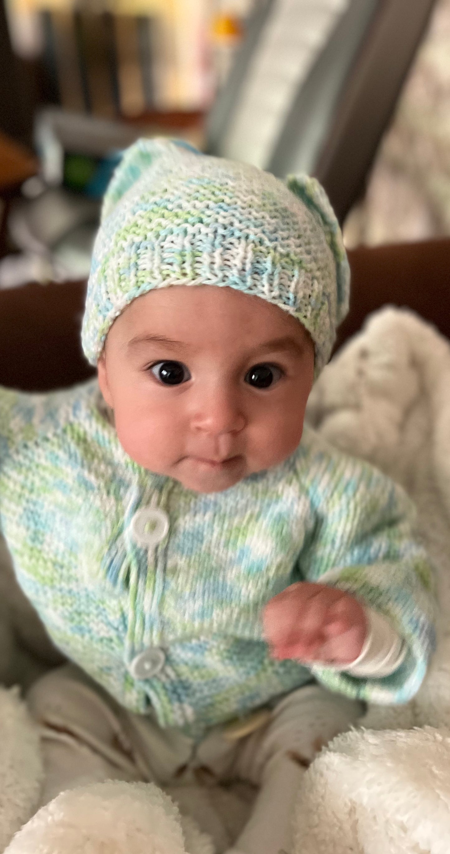 Baby wearing a knitted hat and sweater sitting on a soft surface.