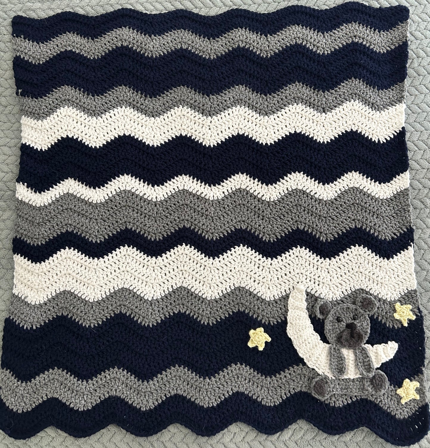 Crocheted blanket with wavy pattern and teddy bear design on a textured surface