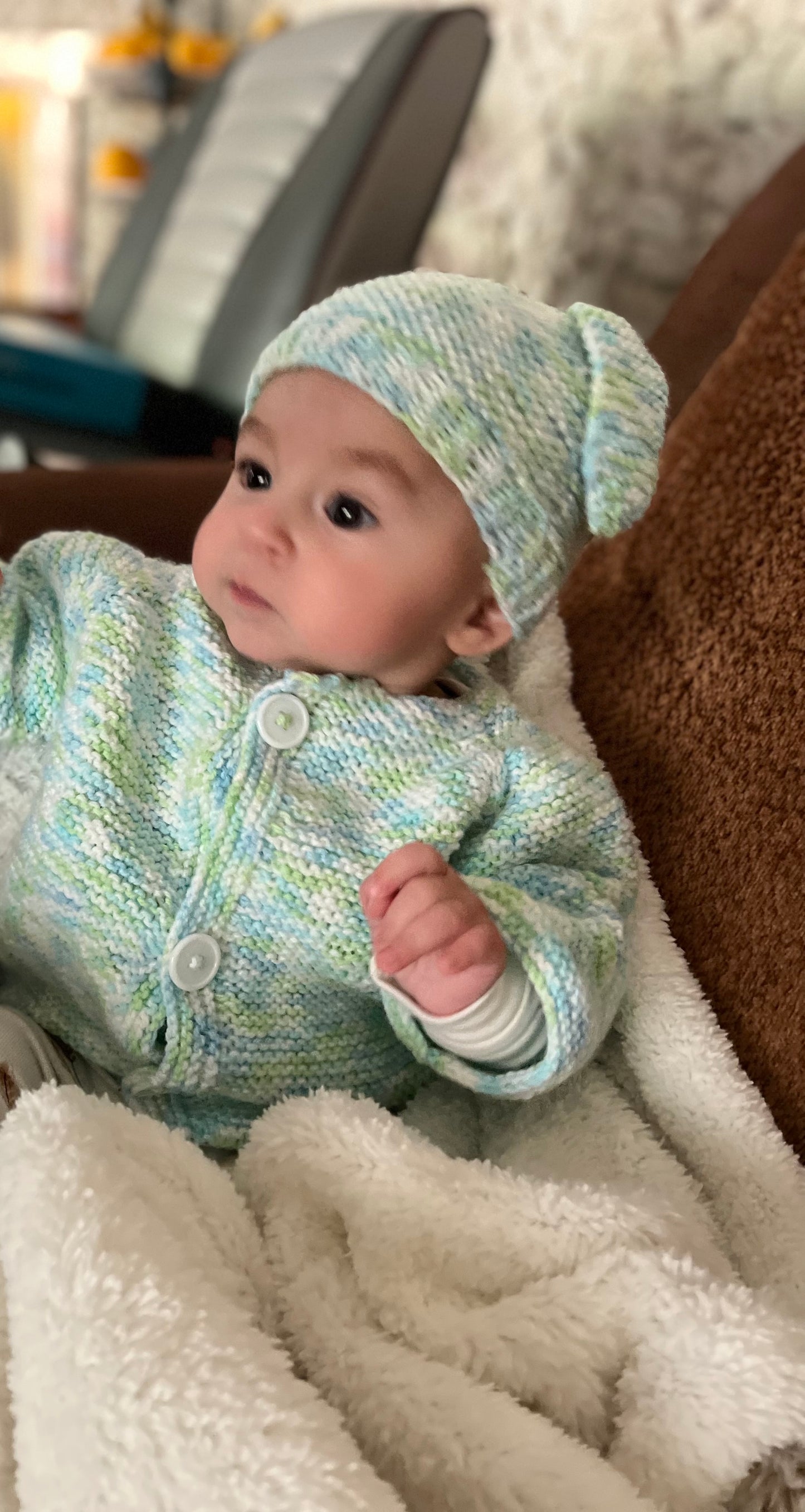 Baby wearing a knitted outfit and hat sitting on a soft surface.