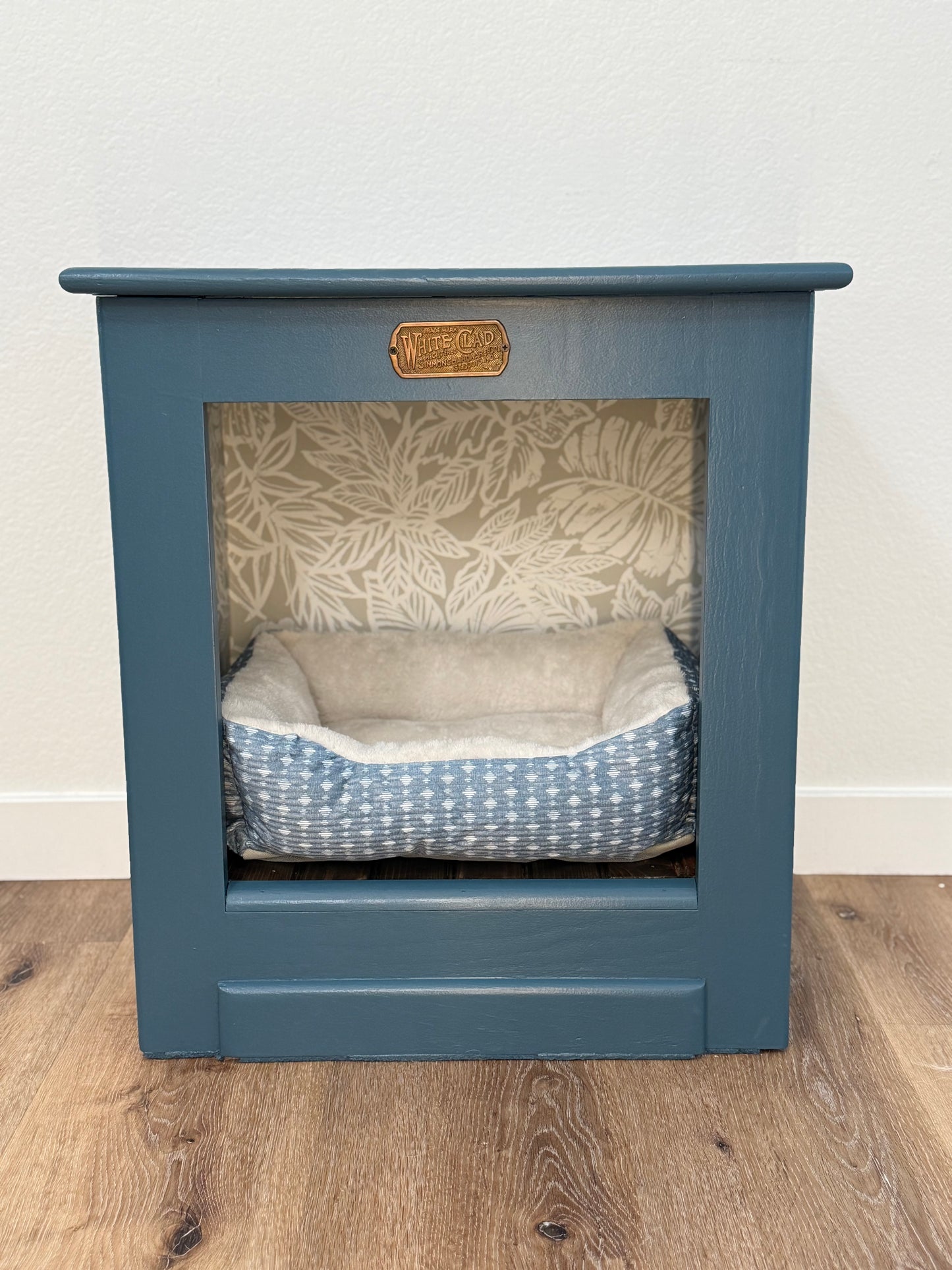 Pet bed inside a blue wooden cabinet with a leaf patterned wallpaper and a wooden floor.