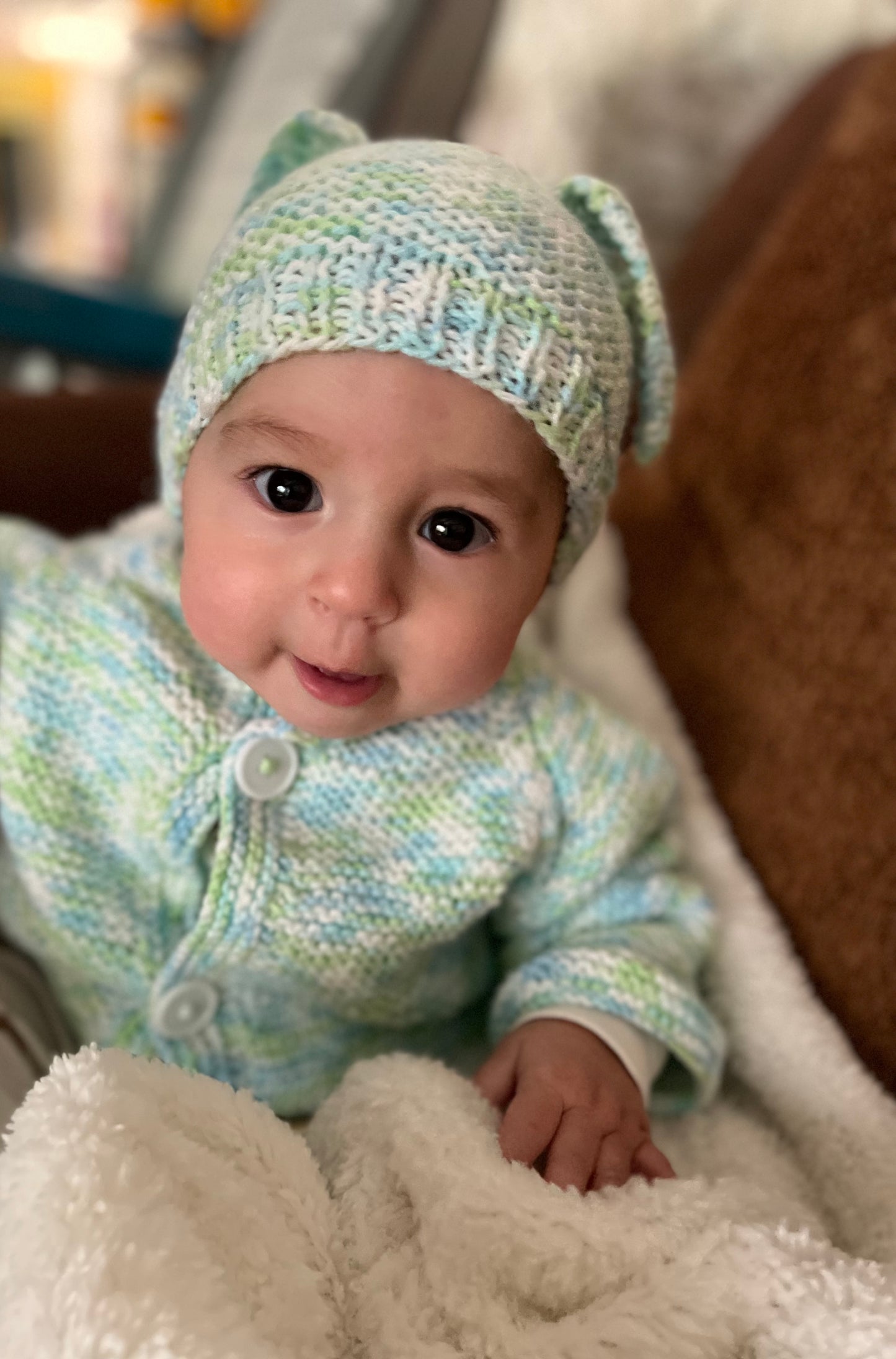 Baby wearing a knitted outfit with a hat featuring ears, sitting on a soft surface.