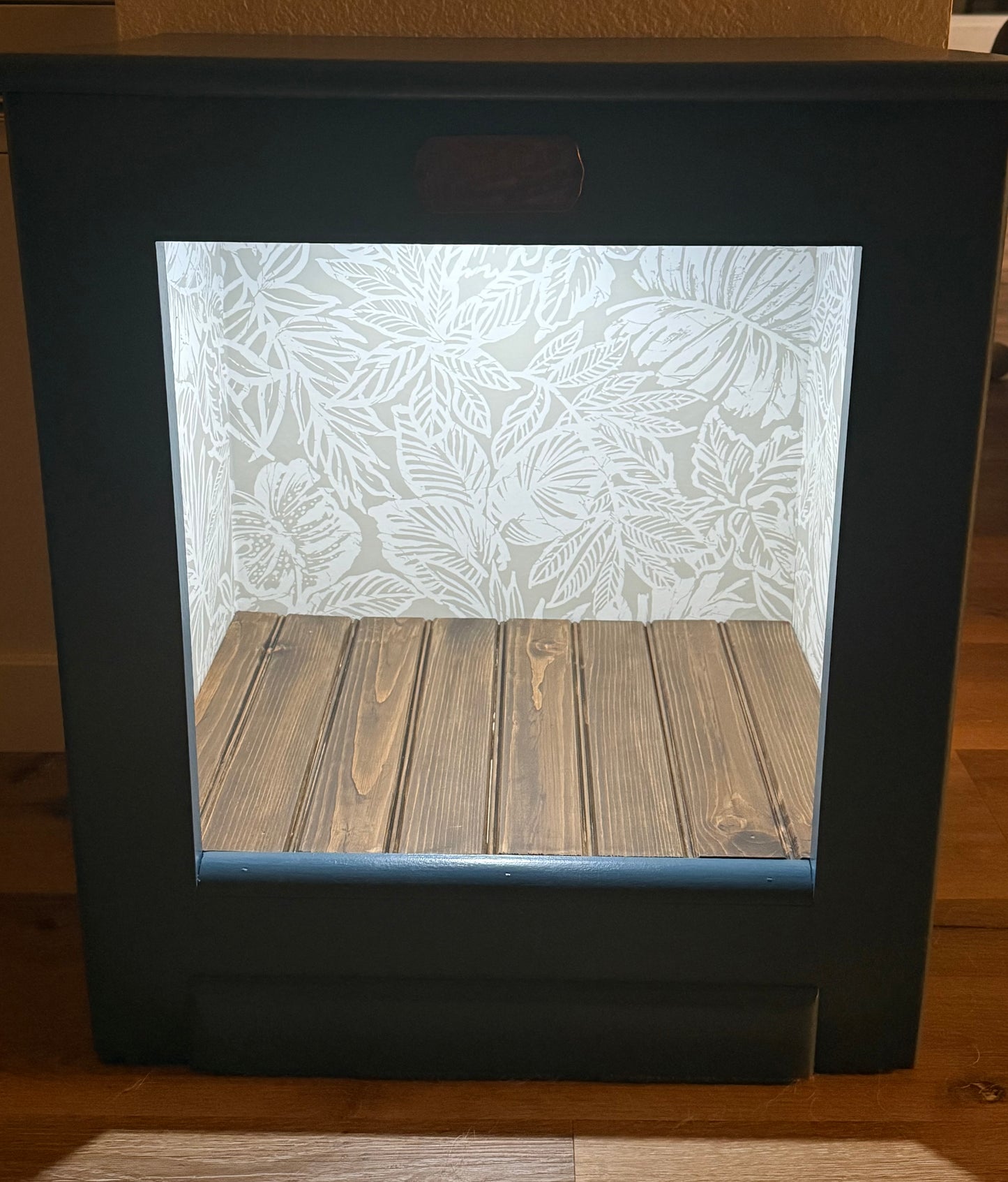Wooden open faced cabinet with a leaf pattern wallpaper on a wooden floor.