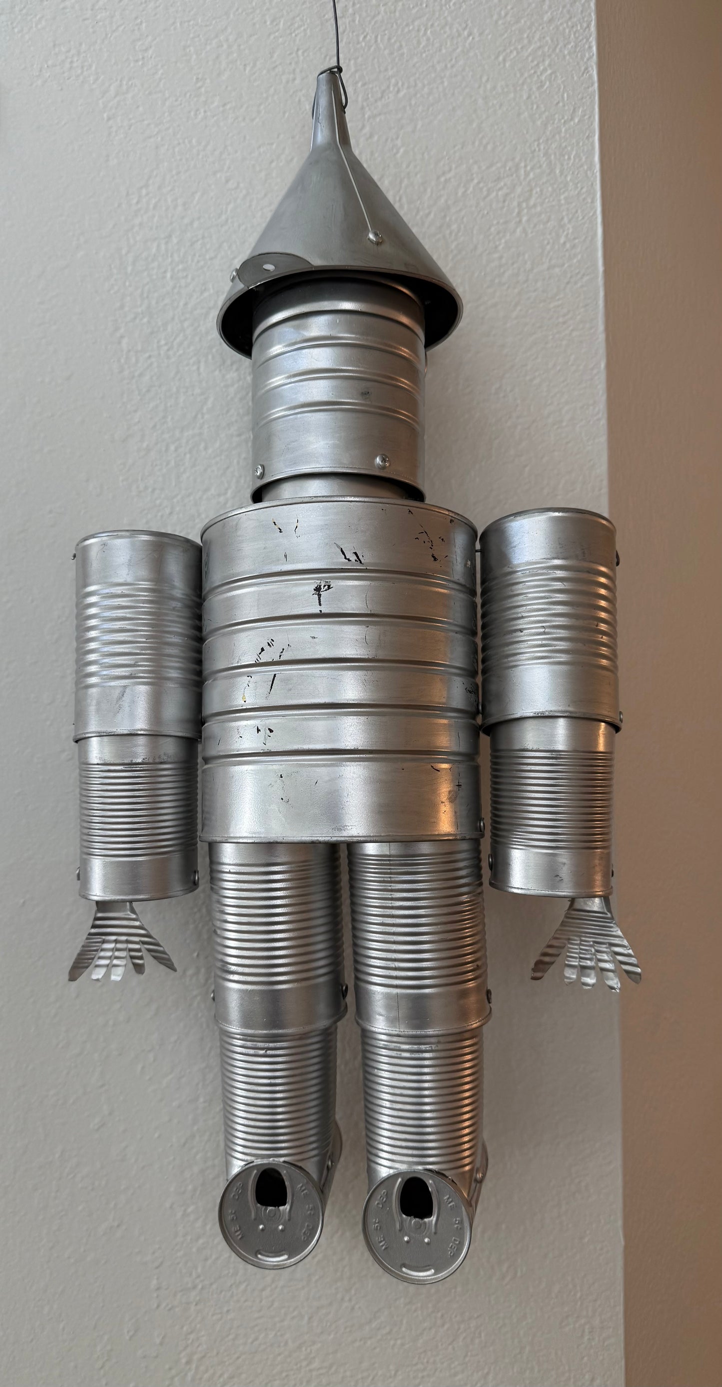 Metallic sculpture resembling the tin man made from tin cans on a plain wall.