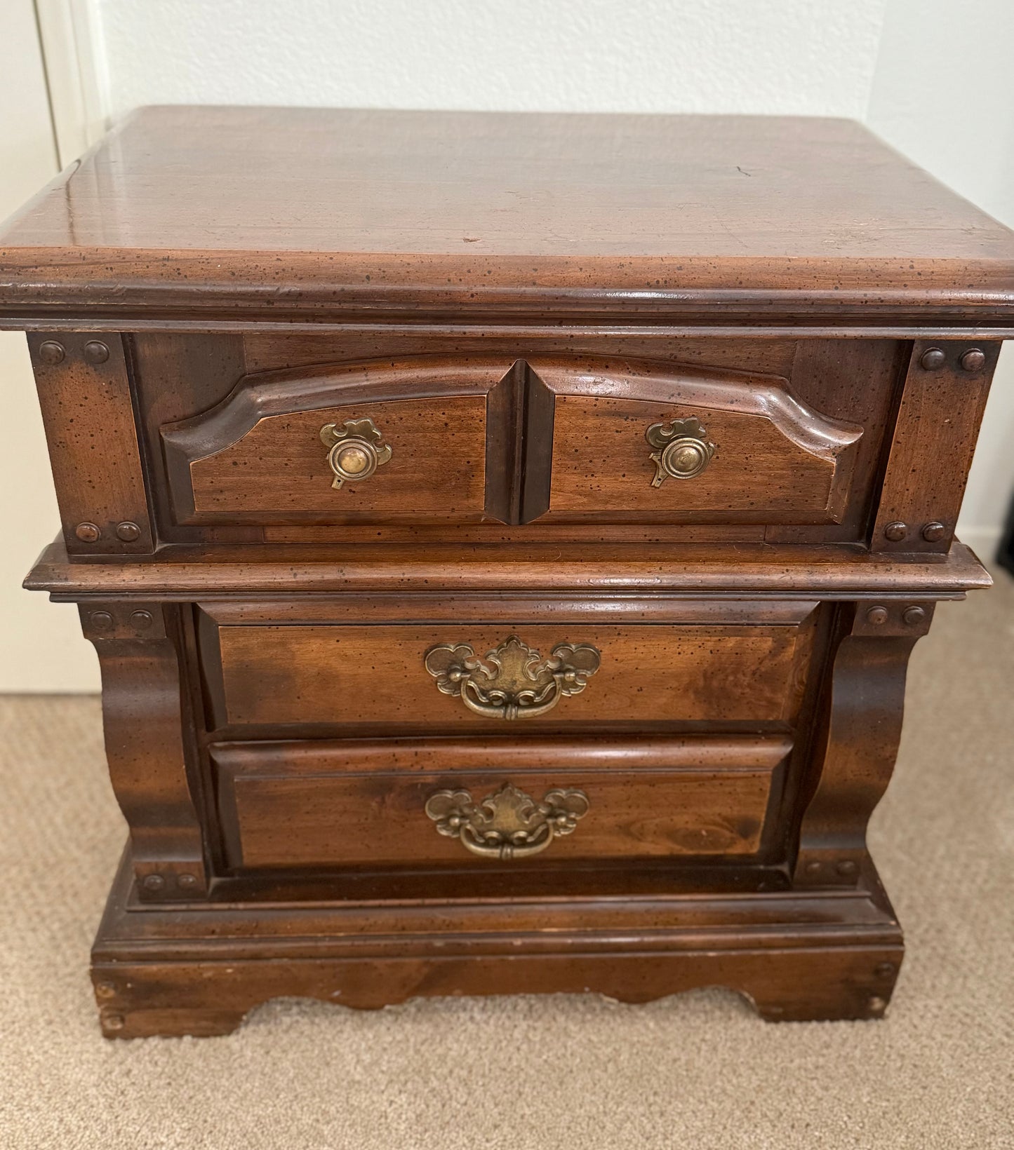 Wooden nightstand with three drawers and brass handles on a carpeted floor.