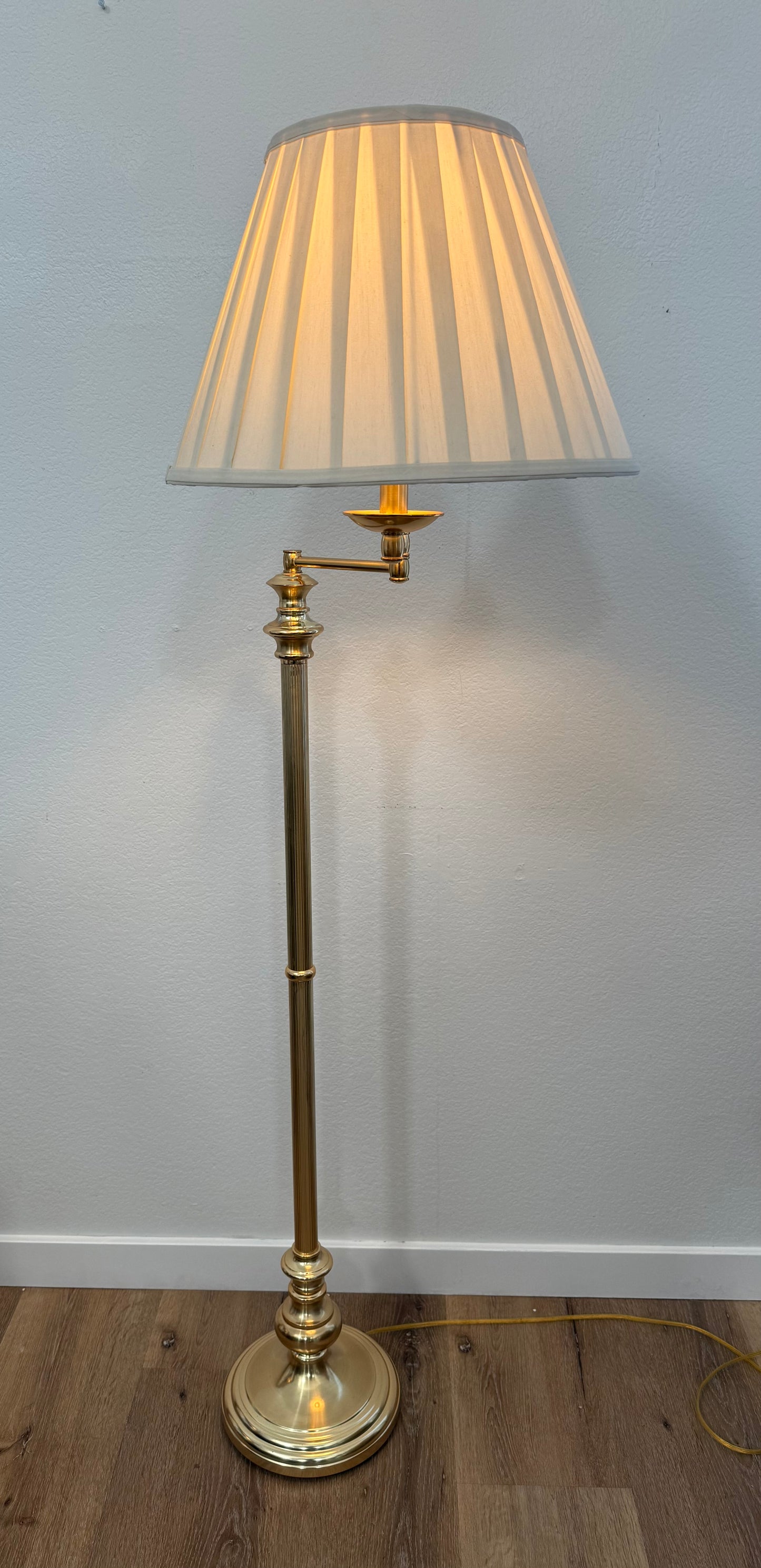 Brass floor lamp with pleated shade against a plain wall.