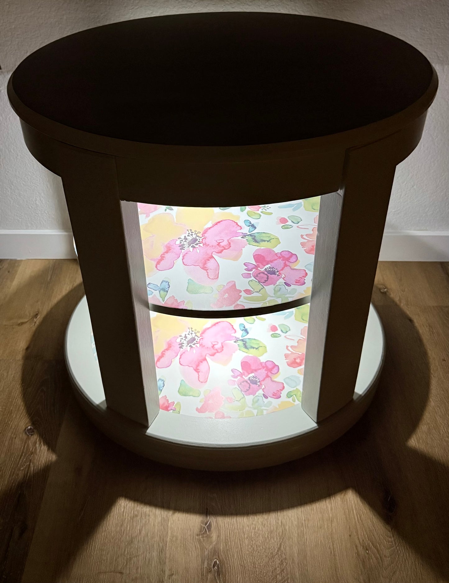 Round side table with floral patterned wallpaper on lighted shelves.