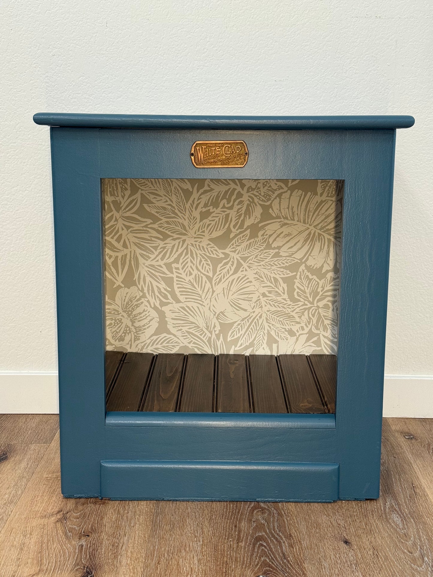 Blue open face White Clad cabinet with interior wallpaper and a wooden floor.