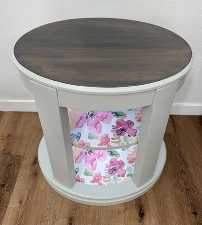 Oval wooden side table with floral patterned shelves on a wooden floor.