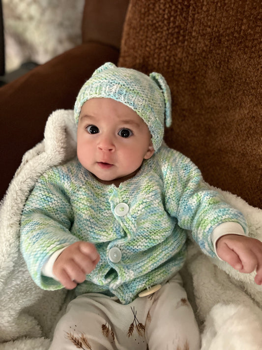 Baby wearing a green knitted sweater and hat sitting on a soft surface.