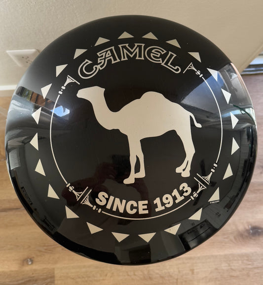 Round black sign with white camel and text on a wooden surface