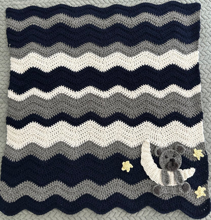 Crocheted blanket with wavy pattern and teddy bear design on a textured surface