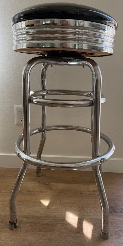 Bar stool with black cushioned seat and chrome frame on a wooden floor.
