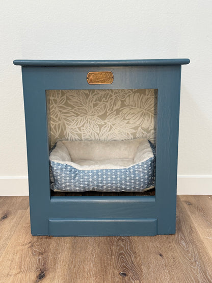 Pet bed inside a blue wooden cabinet with a leaf patterned wallpaper and a wooden floor.
