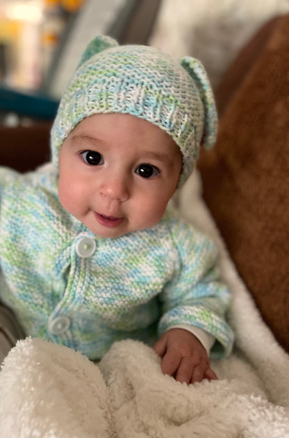 Baby wearing a knitted outfit with a hat featuring ears, sitting on a soft surface.