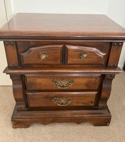 Wooden nightstand with three drawers and brass handles on a carpeted floor.
