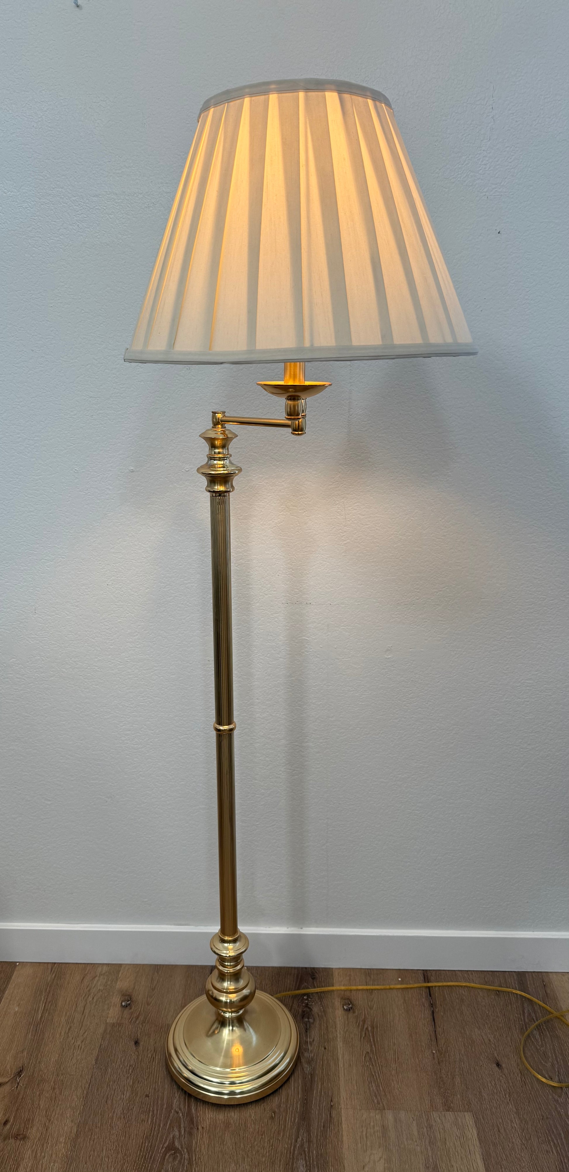 Brass floor lamp with pleated shade against a plain wall.