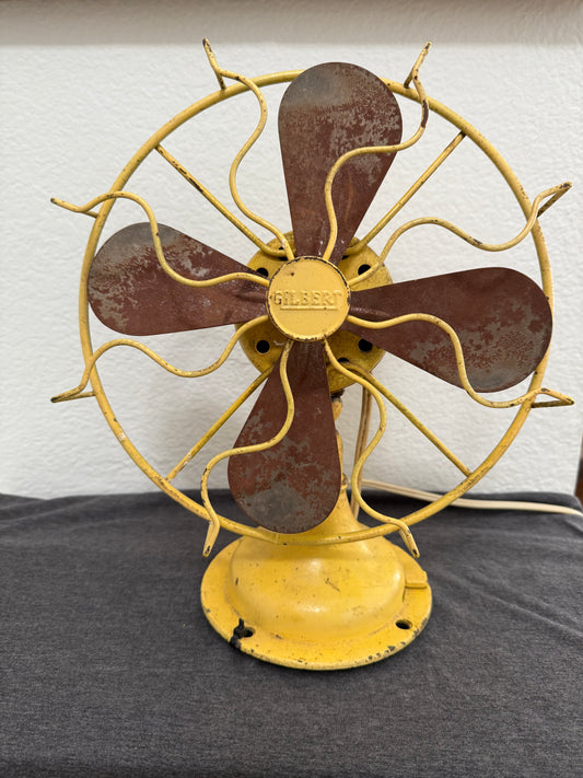 Vintage fan with rusted metal blades and yellow housing on a white wall.