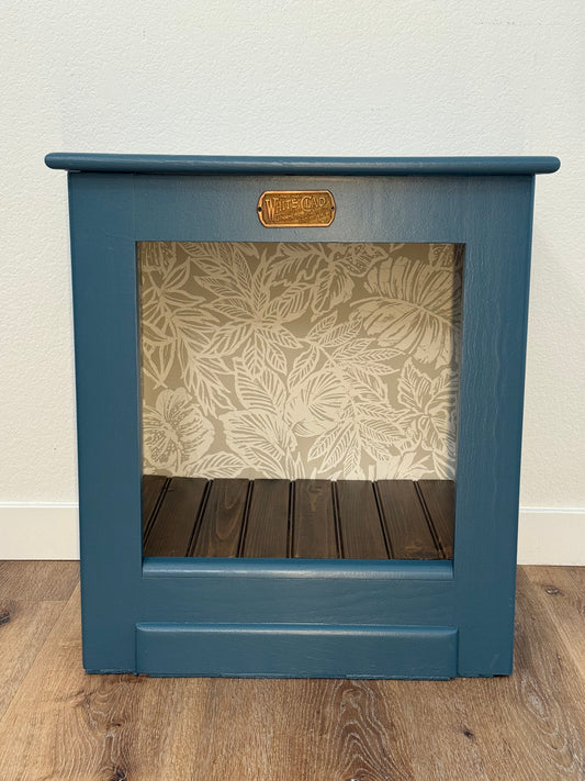 Blue open face White Clad cabinet with interior wallpaper and a wooden floor.